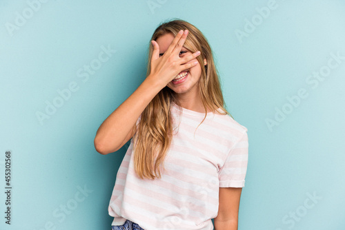 Young caucasian blonde woman isolated on blue background blink at the camera through fingers, embarrassed covering face.