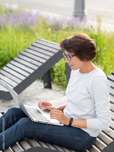 Woman sits with laptop on urban park bench. Freelancer at work. Student learns remotely from outdoors. Modern lifestyle.