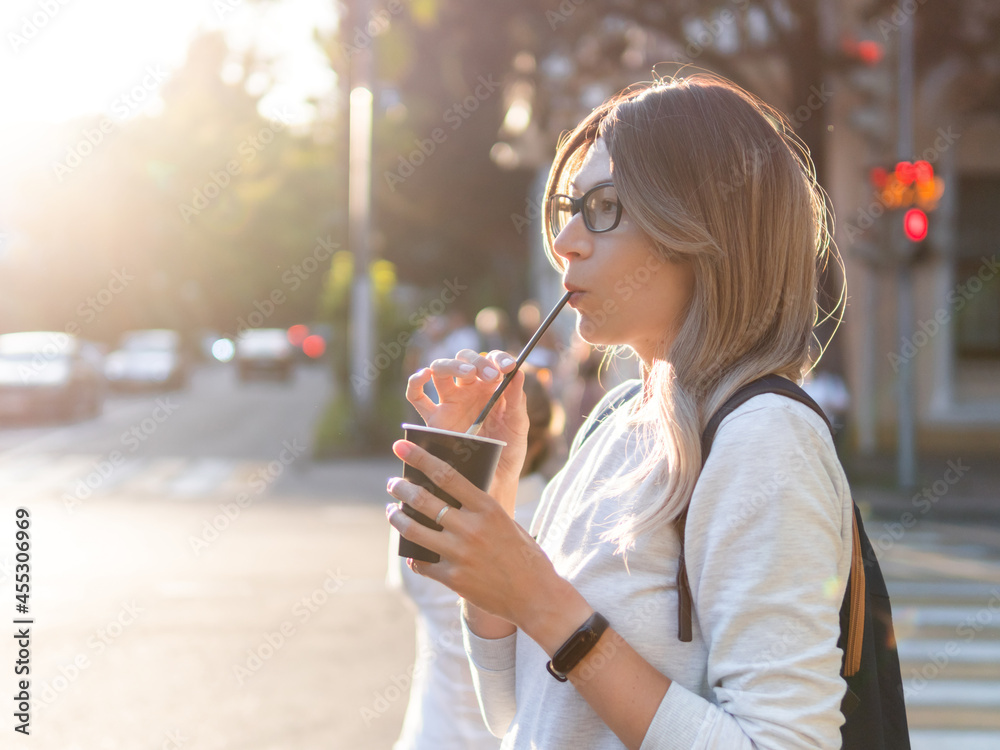 Woman with curly hair and eyeglasses drinks cappuccino with straw. Pretty female waits for green traffic light on pedestrian crossing. Woman with cup of take away coffee. Street life in town.