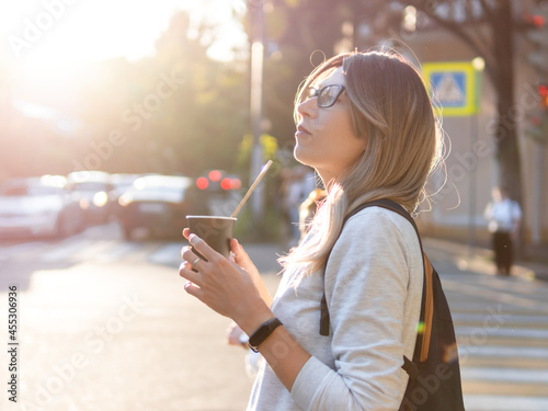 Woman with curly hair and eyeglasses drinks cappuccino with straw. Pretty female waits for green traffic light on pedestrian crossing. Woman with cup of take away coffee. Street life in town.