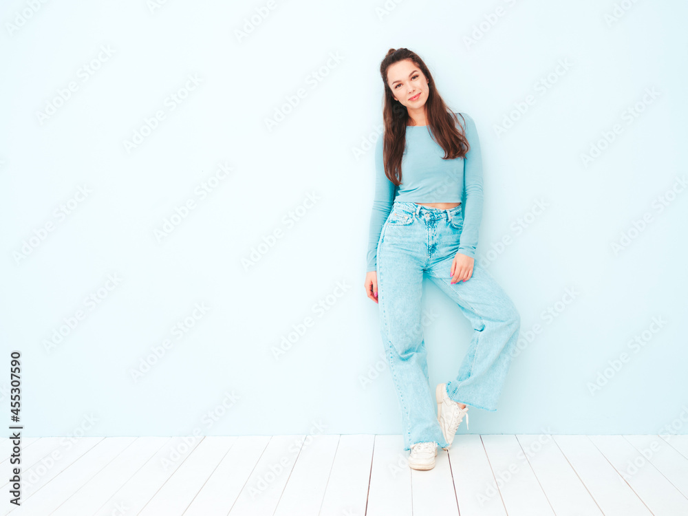Obraz premium Young beautiful smiling female in trendy summer hipster clothes. Sexy carefree woman posing near light blue wall in studio. Positive model having fun indoors. Cheerful and happy