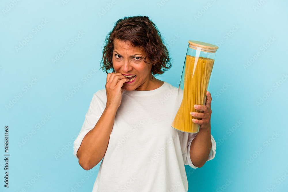 Middle age latin woman holding pasta jar isolated on blue background biting fingernails, nervous and very anxious. Middle age latin woman holding pasta jar isolated on blue background biting fingernails, nervous and very anxious.