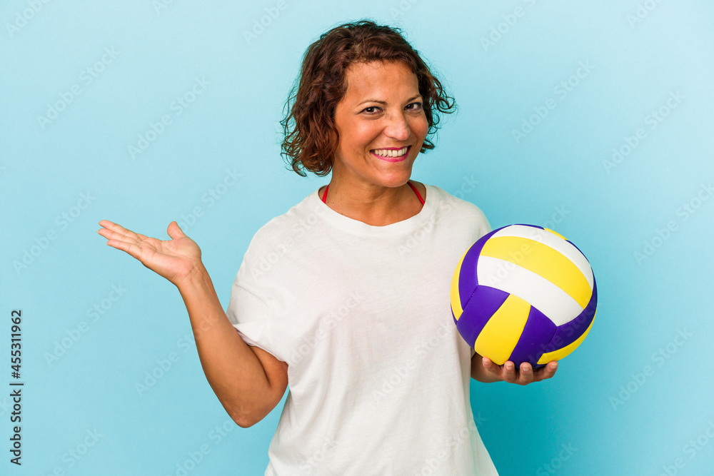 Middle age latin woman playing volleyball isolated on blue background showing a copy space on a palm and holding another hand on waist. Middle age latin woman playing volleyball isolated on blue background showing a copy space on a palm and holding another hand on waist.