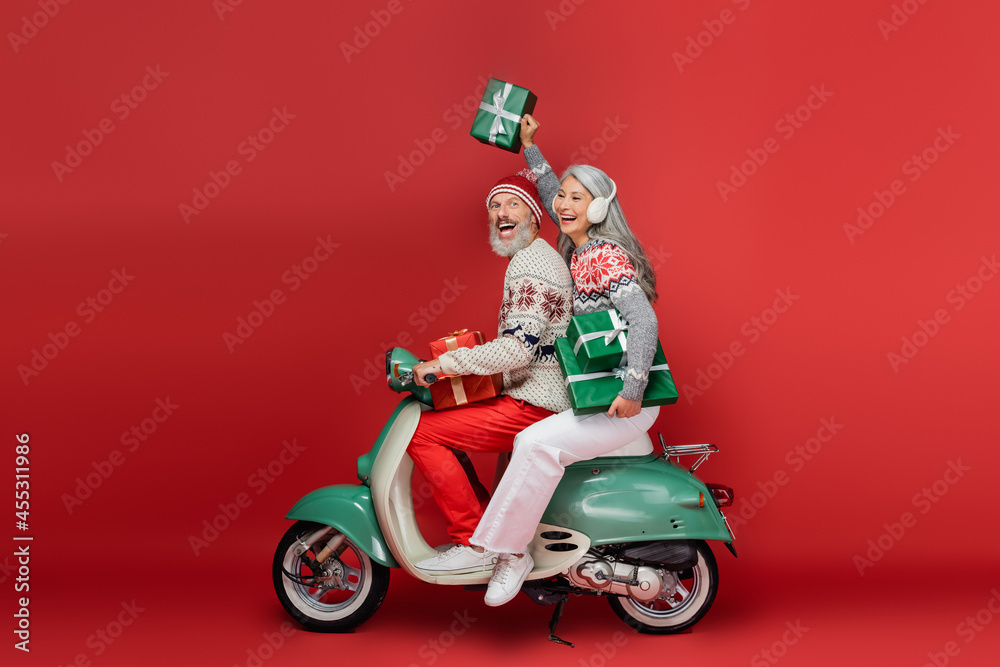 cheerful middle aged asian woman in ear muffs holding christmas presents while riding motor scooter with excited husband on red cheerful middle aged asian woman in ear muffs holding christmas presents while riding motor scooter with excited husband on red