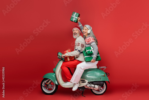 cheerful middle aged asian woman in ear muffs holding christmas presents while riding motor scooter with excited husband on red