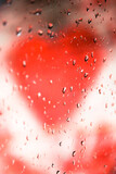 A blurry red heart shape behind glass window with raindrops on a surface. Valentine's Day, unhappy love concept. Gloomy autumn day. Bad weather outdoor, outside. Vertical photo. Love message.