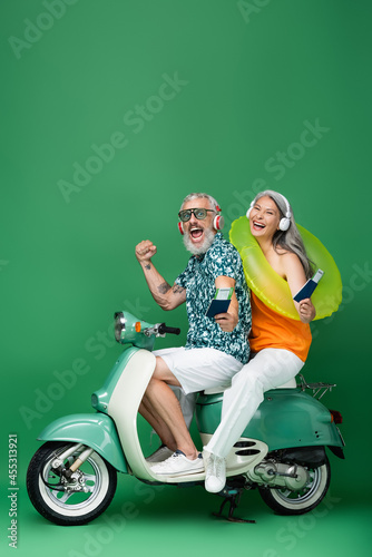 multiethnic and middle aged couple in wireless headphones holding passports with air tickets and riding moped on green