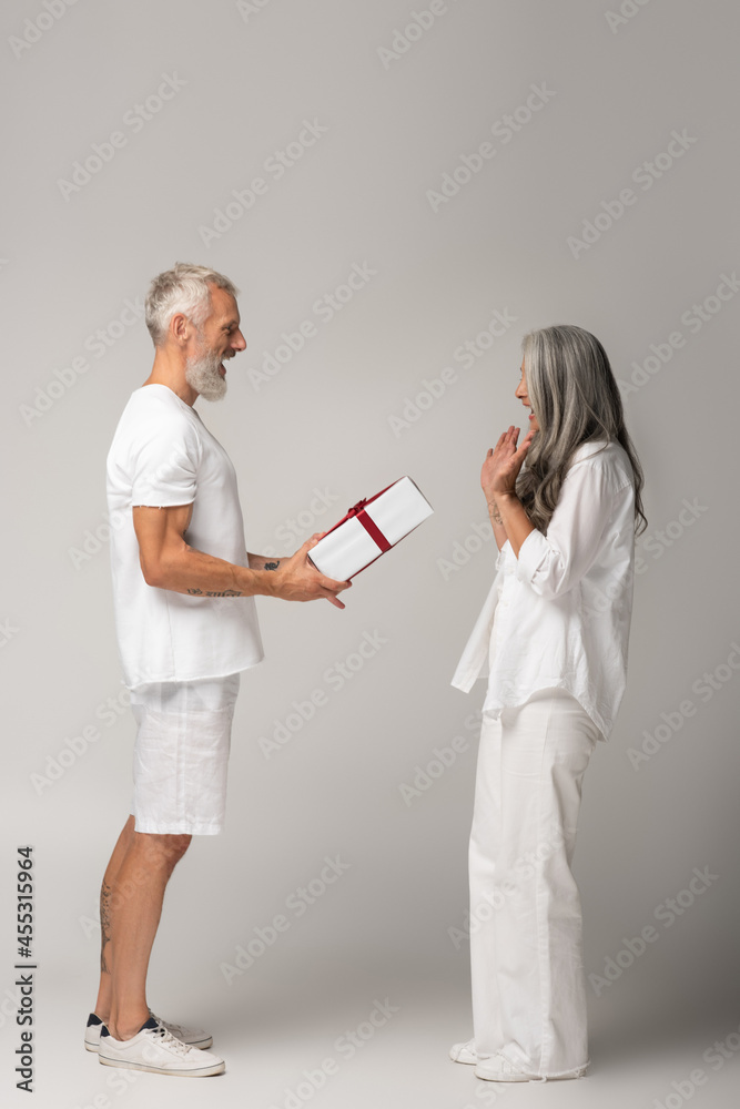 full length of happy middle aged man giving wrapped gift box to amazed asian wife on grey full length of happy middle aged man giving wrapped gift box to amazed asian wife on grey