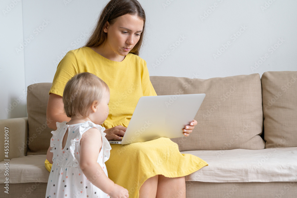 Blond baby in dress standing next to mother sitting on couch and looking at laptop screen. Curious toddler distractioning mother from working at home. Concept of remote work challenges Blond baby in dress standing next to mother sitting on couch and looking at laptop screen. Curious toddler distractioning mother from working at home. Concept of remote work challenges