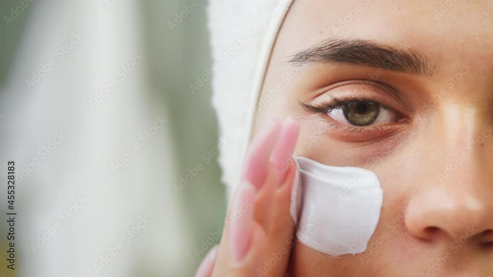 Woman using moisturizing face cream, morning skincare routine. Young female student wearing towel on head looking in mirror in spa salon. Make-up preparation. Half-face portrait. Woman using moisturizing face cream, morning skincare routine. Young female student wearing towel on head looking in mirror in spa salon. Make-up preparation. Half-face portrait.
