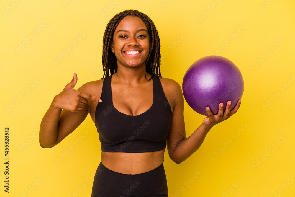 Young african american woman holding a pilates ball isolated on yellow background person pointing by hand to a shirt copy space, proud and confident Young african american woman holding a pilates ball isolated on yellow background person pointing by hand to a shirt copy space, proud and confident
