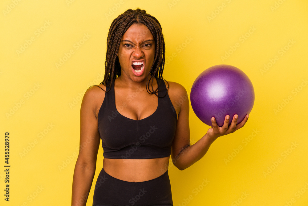 Young african american woman holding a pilates ball isolated on yellow background screaming very angry and aggressive. Young african american woman holding a pilates ball isolated on yellow background screaming very angry and aggressive.