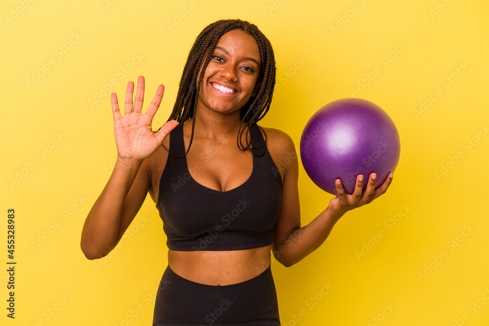 Young african american woman holding a pilates ball isolated on yellow background smiling cheerful showing number five with fingers. Young african american woman holding a pilates ball isolated on yellow background smiling cheerful showing number five with fingers.