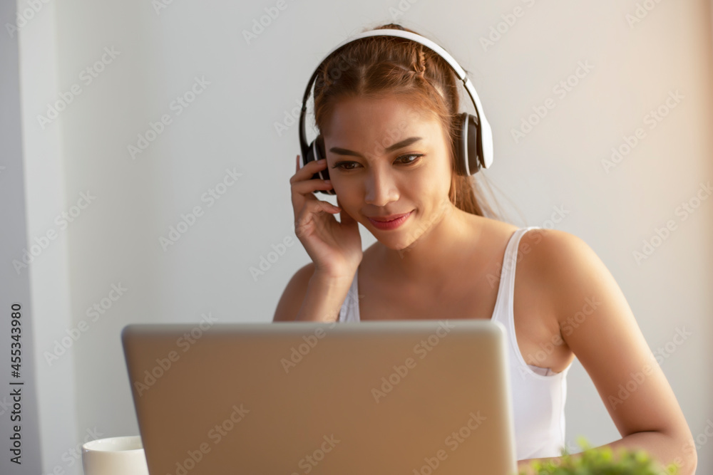 Asian woman wearing headphones using laptop sitting in her bedroom to communicate with friends during the epidemic of COVID-19