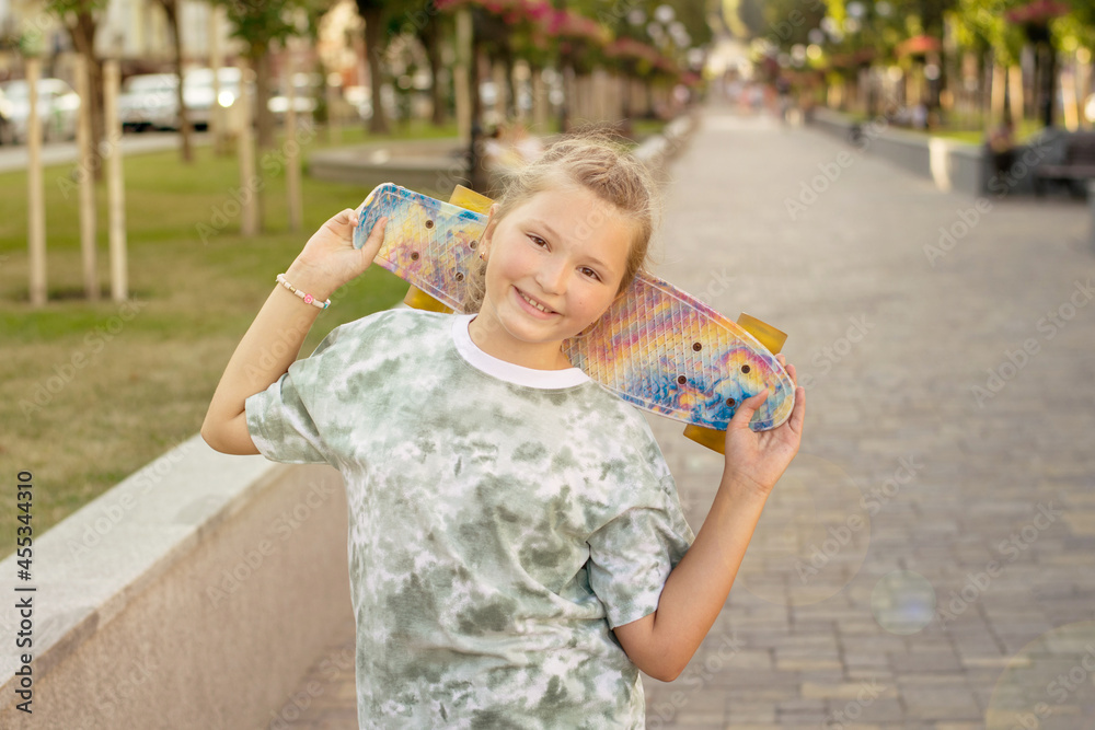 Naklejka premium Cute smiling teenager girl with skateboard in the park on a summer sunny day having fun. Healthy lifestyle and leisure concept for teenagers