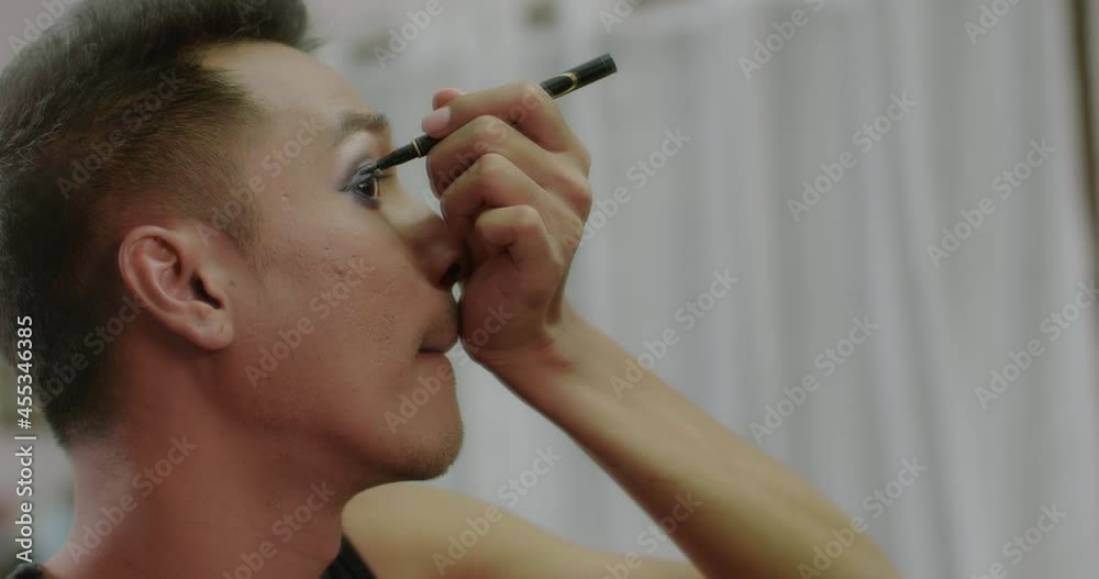 A charming short-haired Asian gay who owns a small beauty salon in the village is writing his eyeliner as he uses his mobile phone to record a video to post on social media. A charming short-haired Asian gay who owns a small beauty salon in the village is writing his eyeliner as he uses his mobile phone to record a video to post on social media.