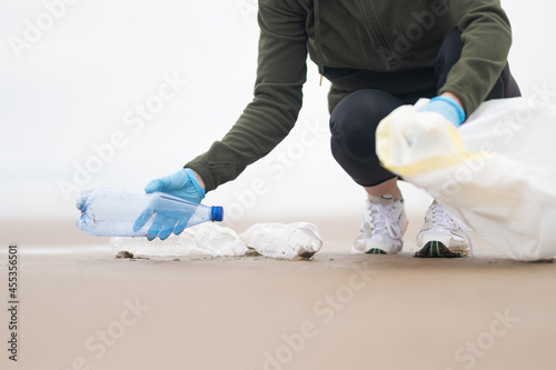 A girl volunteer in blue gloves puts plastic bottles in a large white bag. Cleaning the environment from debris. Environmental and social issues. There is an empty space for insertion.