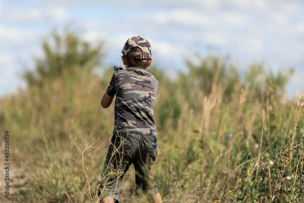 a little boy in a camouflage cap and shorts plays with a gun in the green grass