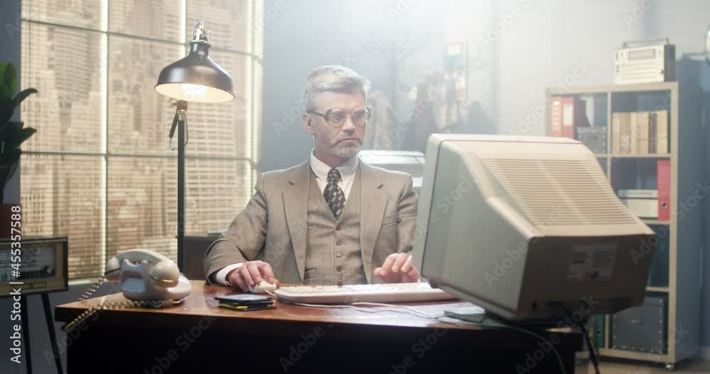 Caucasian male in glasses sitting at the desk in retro room and working on computer. Man programist typing on keyboard and looking seriously. Vintage style of 90's