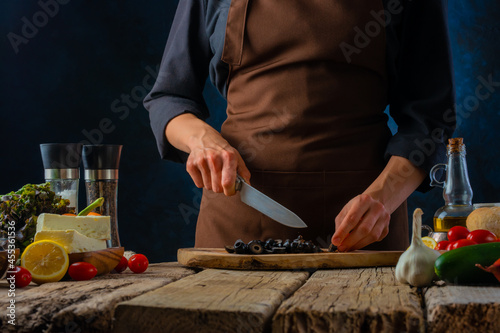 The chef cuts the olives for the classic Greek vitamin salad. Feta cheese  garlic  tomatoes and other ingredients. Wooden texture. Dark background. Restaurant  hotel  step by step recipe.