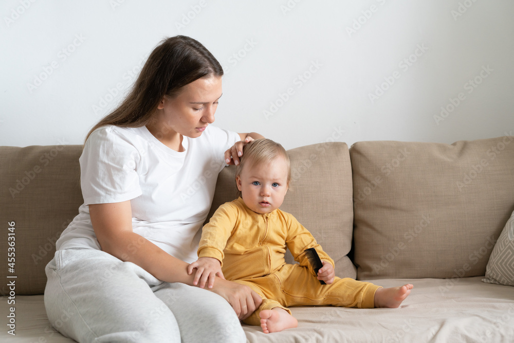 Horizontal portrait of young mother in casual sitting on sofa with adorable blond blue-eyed girl and grooming baby hair. Concept of mother care. Copy space Horizontal portrait of young mother in casual sitting on sofa with adorable blond blue-eyed girl and grooming baby hair. Concept of mother care. Copy space