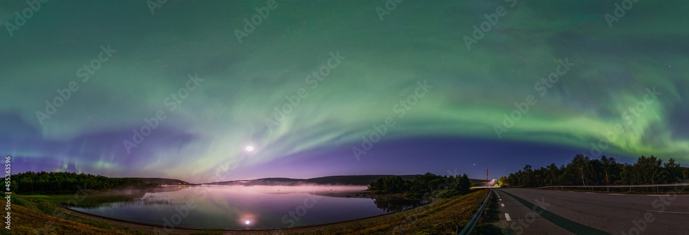 Obraz premium Panoramic Aurora borealis, Northern green lights with full moon and stars in the night sky over mountain lake, mirrored reflection in water, night mist. Night road, Joesjo, Lappland, Sweden