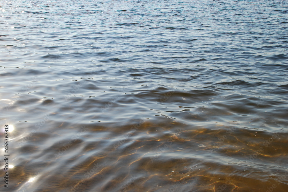 Sandy river bed under the sun. Summer sunny day the surface of the water with small waves, the water is clear, transparent with a brown tint from the yellow-brown sand at the bottom of the reservoir. Sandy river bed under the sun. Summer sunny day the surface of the water with small waves, the water is clear, transparent with a brown tint from the yellow-brown sand at the bottom of the reservoir.