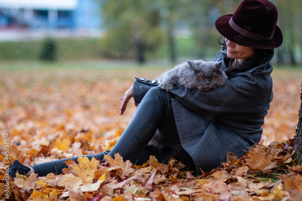 A girl in a hat sits on yellow autumn foliage under a tree and holds a cat in her arms. Autumn mood. A girl in a hat sits on yellow autumn foliage under a tree and holds a cat in her arms. Autumn mood.
