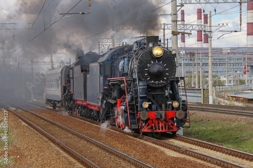 A black steam locomotive with a passenger carriage rides on the railway, releasing puffs of smoke