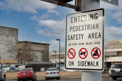 A close up view of a posted sign with graphics and text that reads "Entering Pedestrian Safety Area" with out of focus city background, blue sky and white clouds 