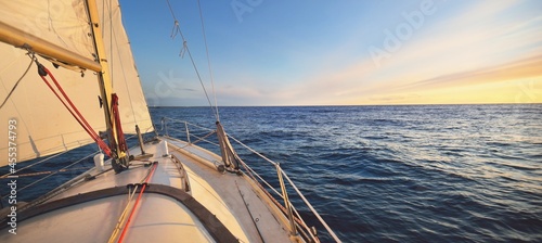 Yacht sailing in an open sea at sunset. Close-up view of the deck, mast and sails. Clear sky after the rain, glowing clouds, golden sunlight. Panoramic seascape photo