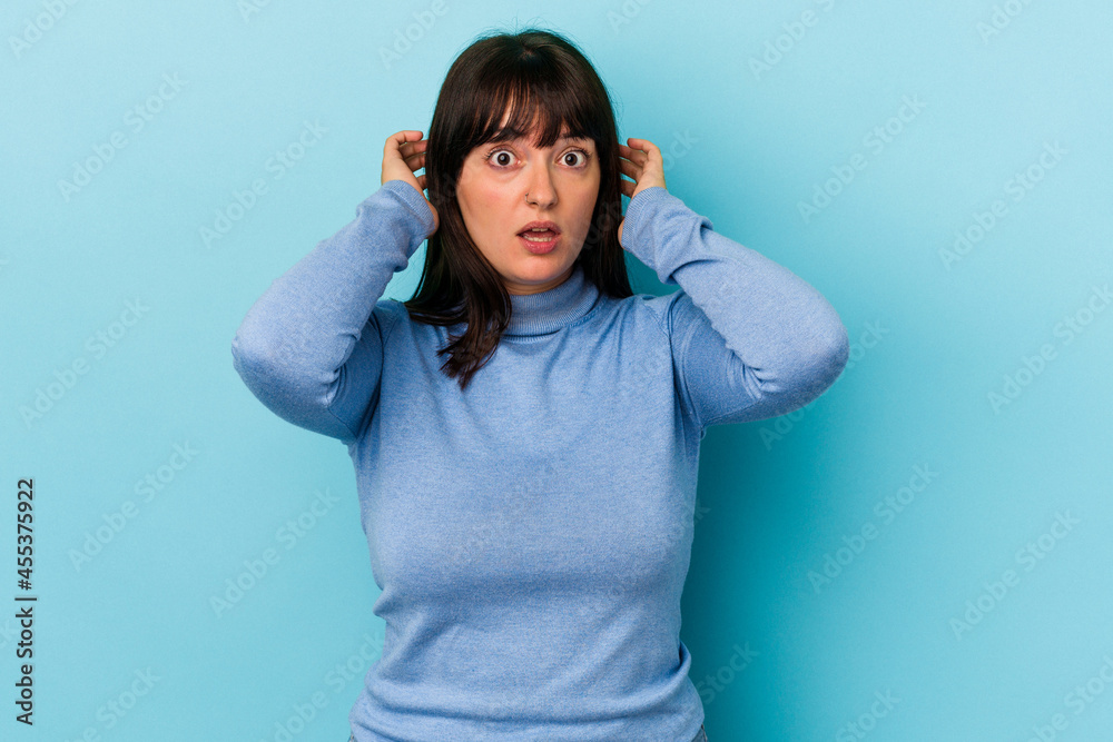Young curvy caucasian woman isolated on blue background covering ears with hands trying not to hear too loud sound.