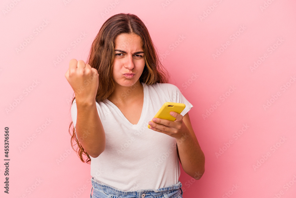 Young caucasian woman using mobile phone isolated on pink background  showing fist to camera, aggressive facial expression.
