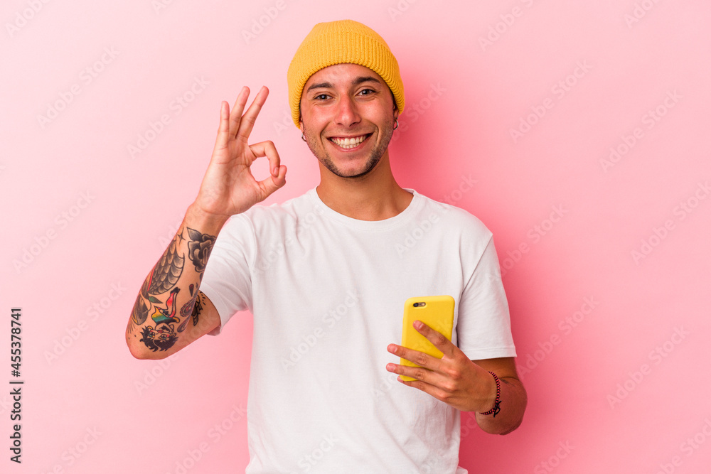 Young caucasian man holding mobile phone isolated on pink background cheerful and confident showing ok gesture. Young caucasian man holding mobile phone isolated on pink background cheerful and confident showing ok gesture.