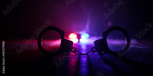 Police raid at night and you are under arrest concept. Silhouette of handcuffs with police car on backside. Image with the flashing red and blue police lights at foggy background.