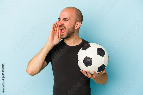 Young business bald man playing football isolated on blue background shouting and holding palm near opened mouth.