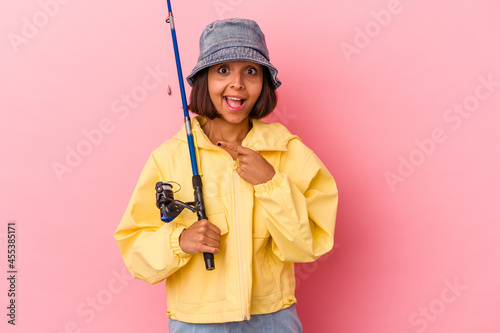 Young mixed race woman practicing fishing isolated on pink background smiling and pointing aside, showing something at blank space. © Asier