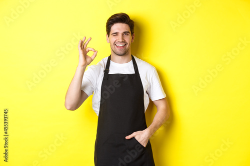 Billede på lærred Confident and handsome waiter showing ok sign, wearing black apron and standing