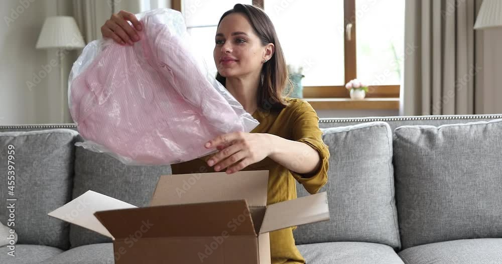 Satisfied of e-commerce online webstore retail services, young woman sit on sofa at home open parcel box, unpack fashion clothes smiles enjoy quality and express delivery e-services. Ecommerce concept