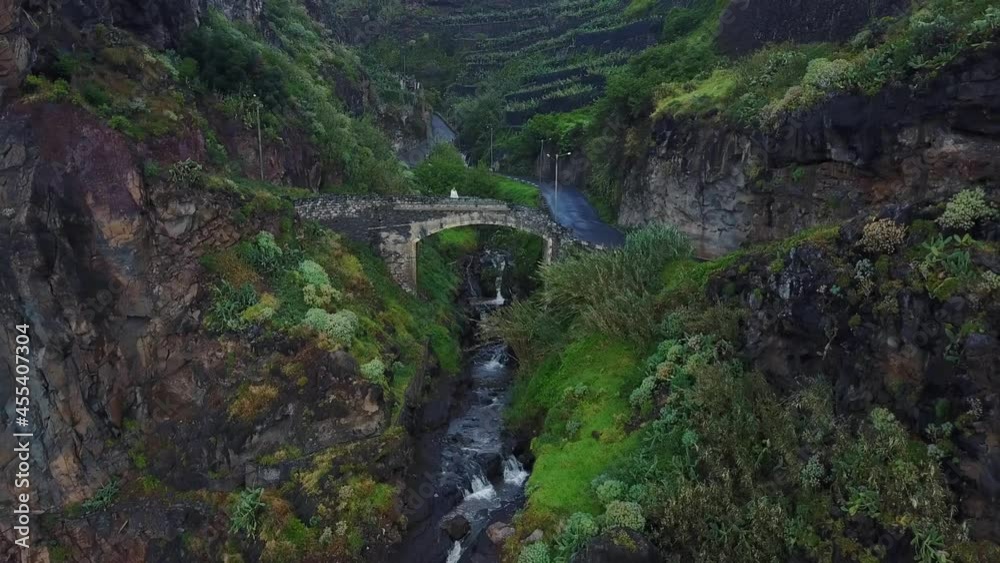 Girl On Ancient Cobblestone Bridge Over Stream Flowing From A Waterfall Cliff to the Atlantic Ocean In Madeira, Portugal. - aerial pullback Girl On Ancient Cobblestone Bridge Over Stream Flowing From A Waterfall Cliff to the Atlantic Ocean In Madeira, Portugal. - aerial pullback