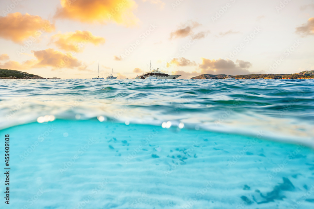 Obraz premium (Selective focus) Split shot, over under picture. Half underwater half sky with a luxury yacht sailing on a turquoise water during a stunning sunrise. Grande Pevero Beach, Sardinia, Italy...