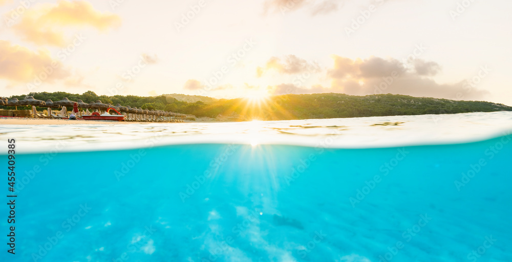(Selective focus) Split shot, over under picture. Crystal clear, turquoise water and some beach umbrellas illuminated during a stunning sunrise. Grande Pevero Beach, Sardinia, Italy.