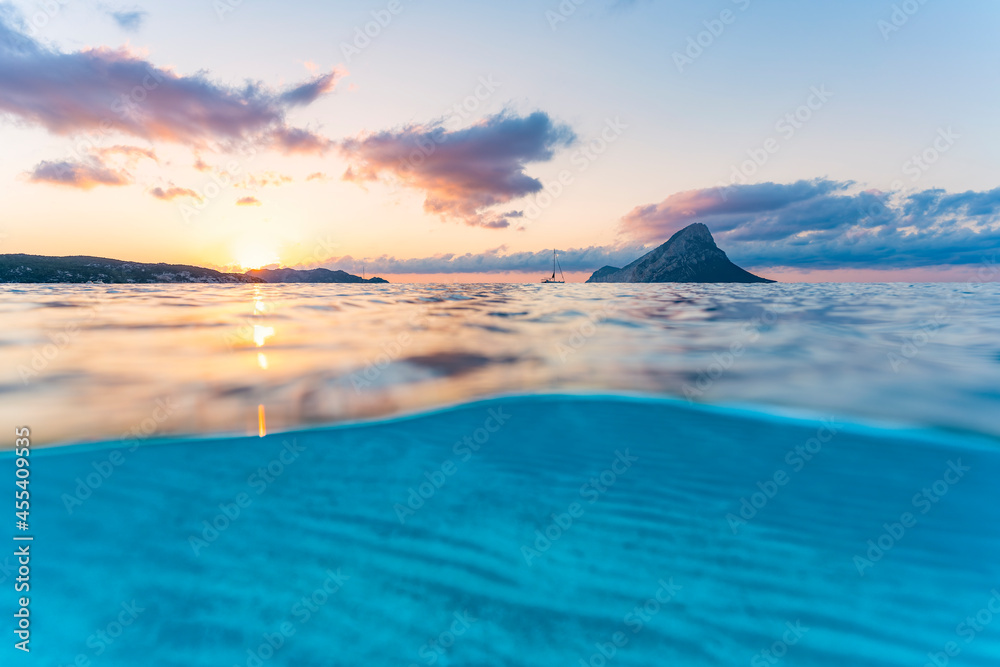 (Selective focus) Split-shot, over-under shot. Turquoise water in the foreground with Tavolara Island on the water surface during a stunning sunrise. Porto Taverna, Sardinia, Italy.