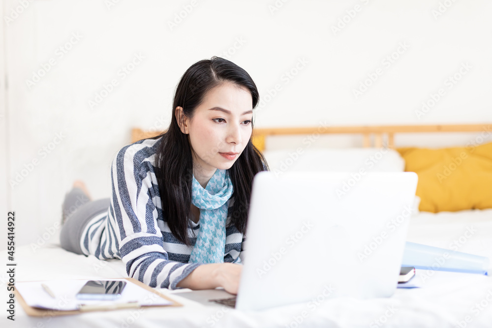 Asian woman typing on her laptop computer while laying in bed at home. Concept of work from home, freelance, free time and startup business Asian woman typing on her laptop computer while laying in bed at home. Concept of work from home, freelance, free time and startup business