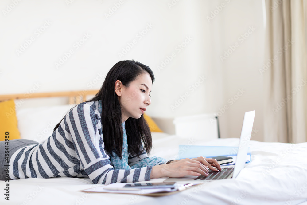 Stressed Asian woman working on her laptop computer while laying in bed at home. Concept of work from home overtime Stressed Asian woman working on her laptop computer while laying in bed at home. Concept of work from home overtime