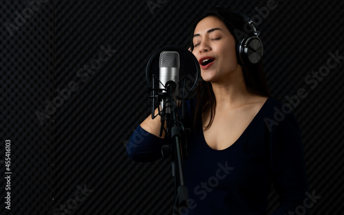 Happy Asain woman as professional singer catching earphone, smile, enjoy practicing attractive voice and song singing performance to microphone in dark sound record room of music studio
