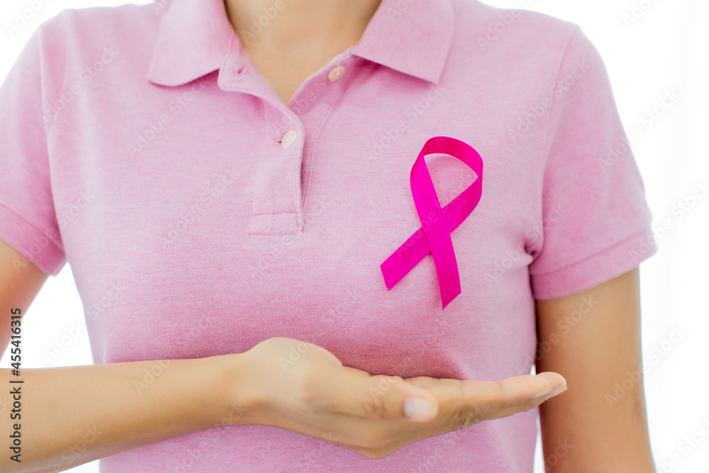 Unrecognizable patient shows hand under breast on pink shirt with ribbon symbol on chest representing cancer awareness campaign. Unrecognizable patient shows hand under breast on pink shirt with ribbon symbol on chest representing cancer awareness campaign.