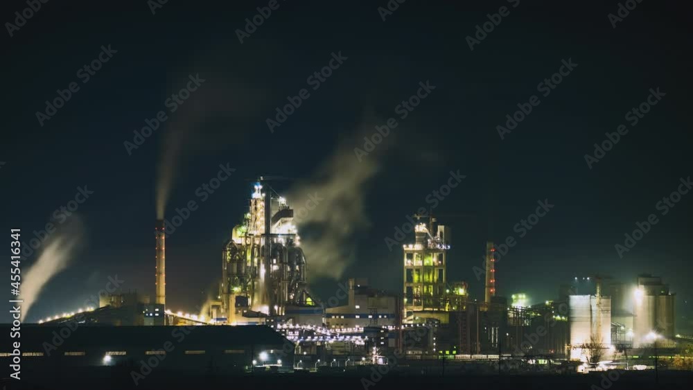 High concrete structure of cement factory with tower crane and smoking chimney at night. Industrial production and air pollution concept.
