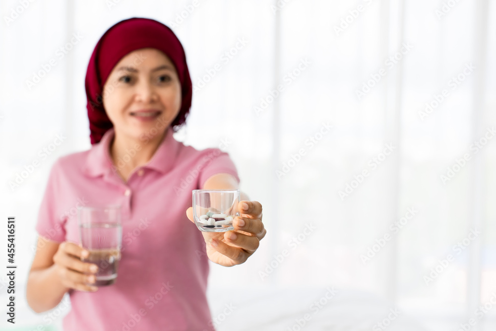 Medicine capsule prepared in clear pharmacy glass on hand of smiling Asian woman on pink shirt and red scarf, hospital patient of breast cancer, before drinking with water to cure disease Medicine capsule prepared in clear pharmacy glass on hand of smiling Asian woman on pink shirt and red scarf, hospital patient of breast cancer, before drinking with water to cure disease