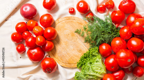Round cutting wooden board, around lies a mountain of red tomatoes, leaves of greenery, dill.